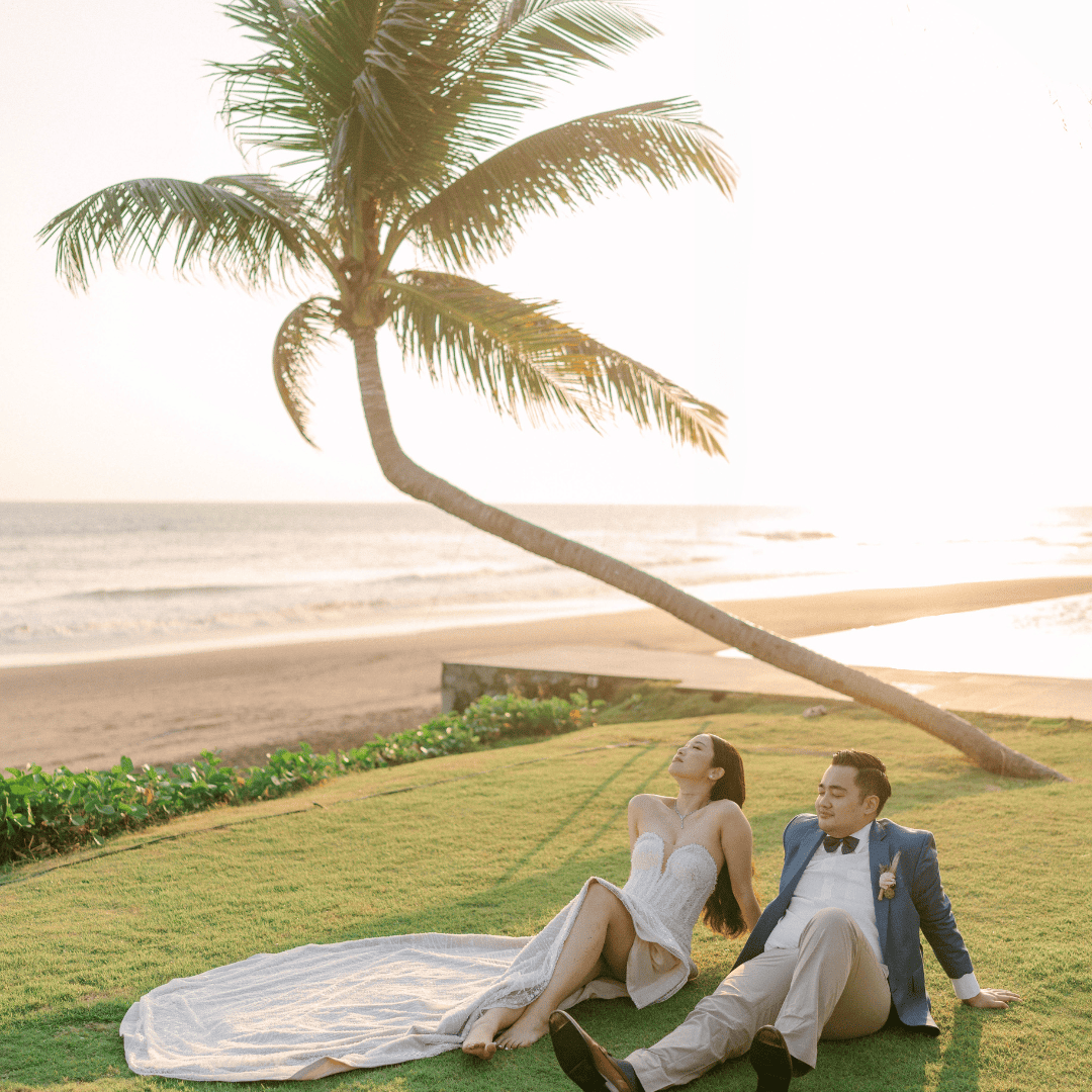 Bride and groom relaxing on grass near beach with a leaning palm tree at sunset.