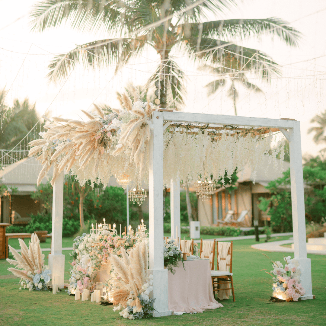 Outdoor wedding reception setup with a white wooden pergola decorated with hanging flowers and chandeliers.