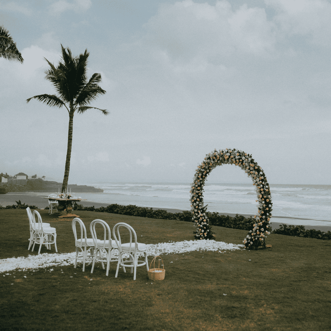 Outdoor wedding setup on grassy area by the ocean with white chairs, flower petal aisle, and floral arch.