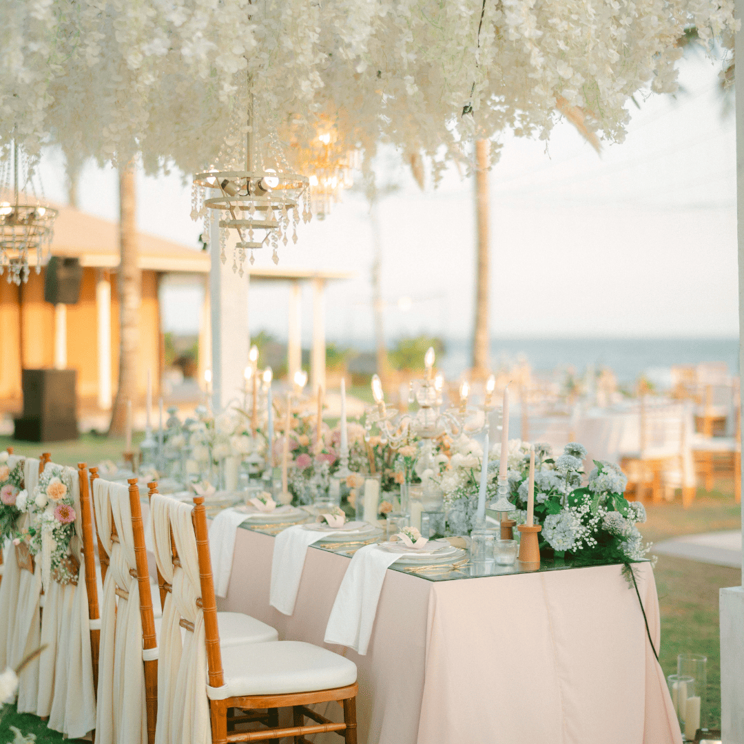 Elegant outdoor dining table with white flowers, candles, and wooden chairs under hanging white floral decorations.