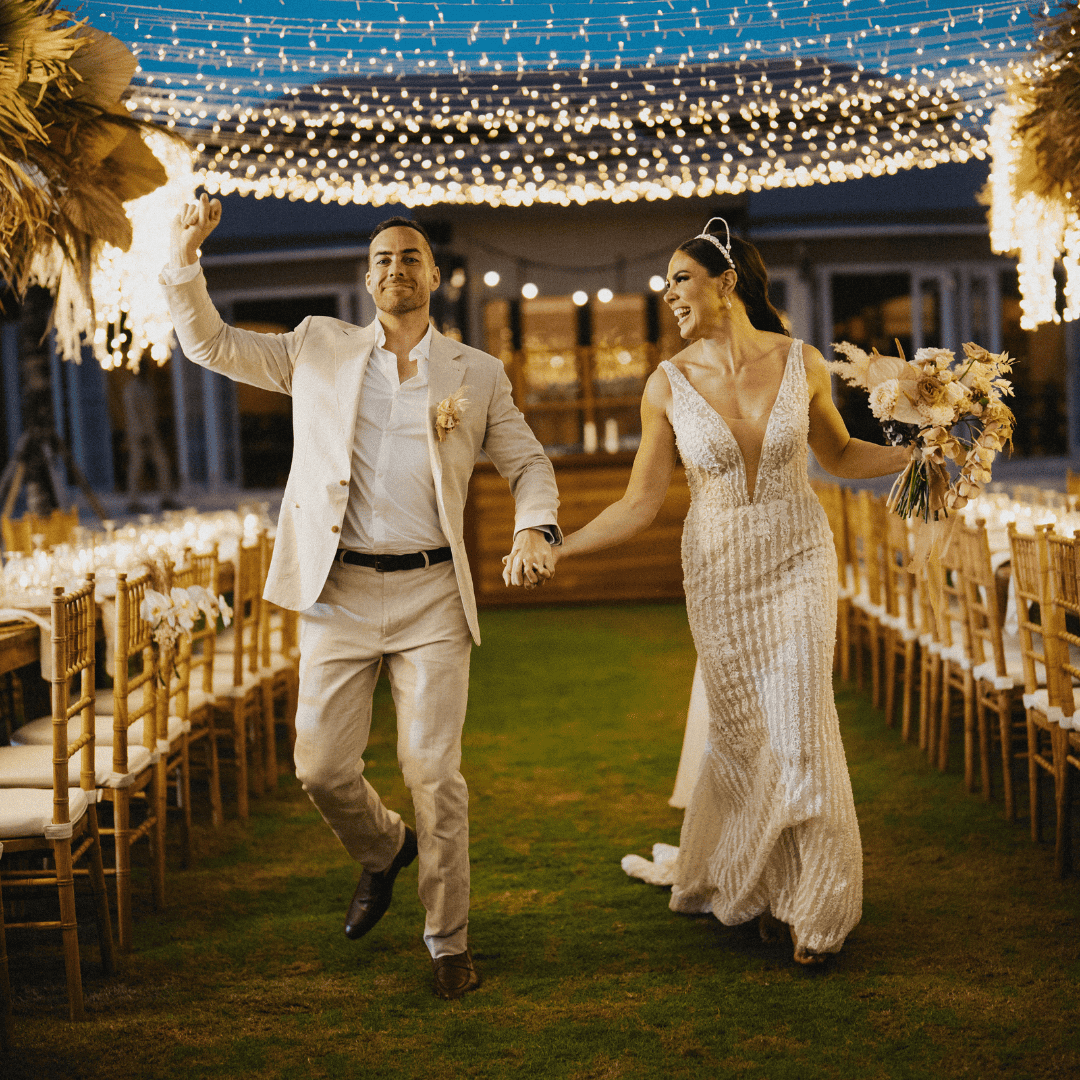 Bride and groom holding hands and smiling under string lights at outdoor wedding reception.