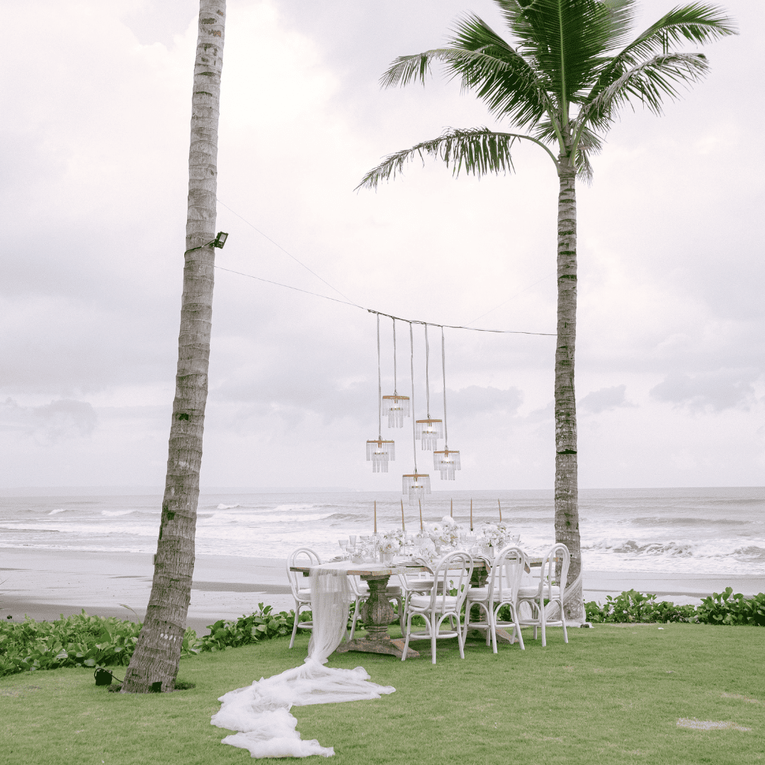 Elegant white table setting with floral centerpieces and candles on grass between two palm trees by the ocean.