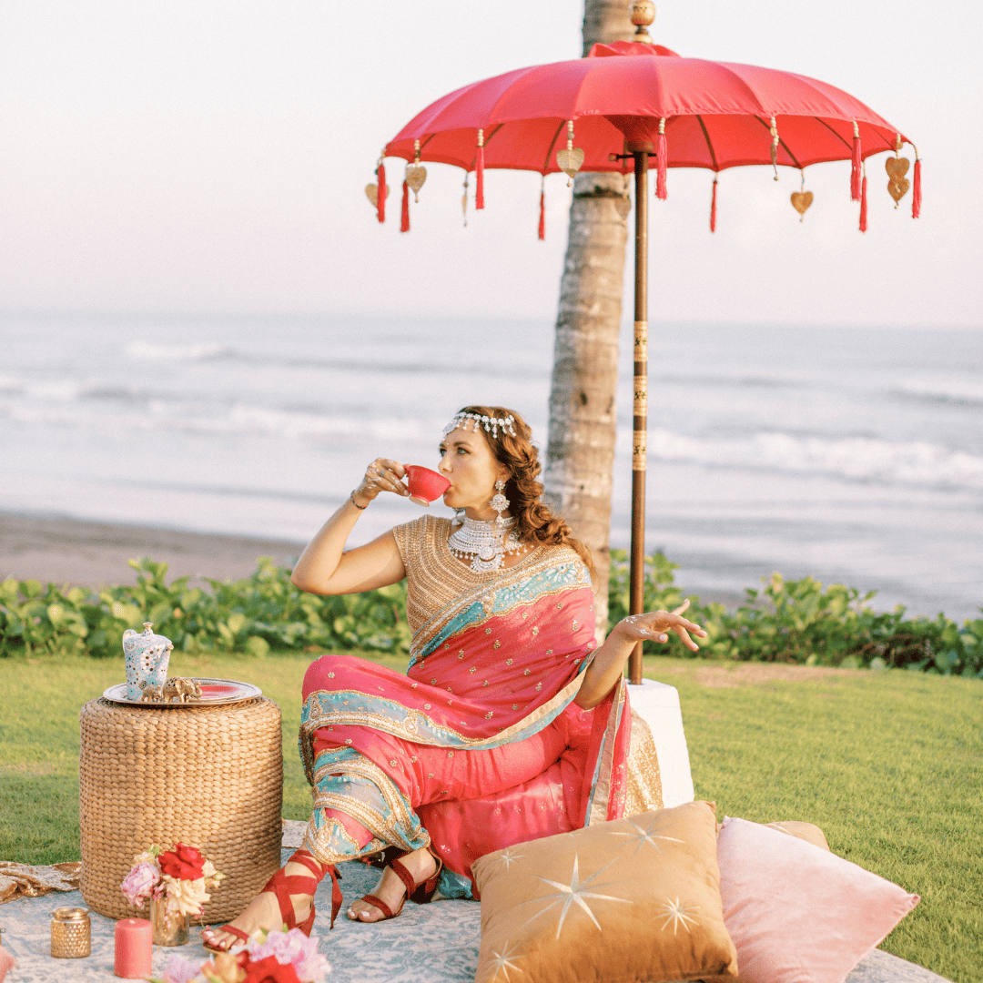 Woman in a pink and blue saree drinking tea under a red umbrella by the ocean.