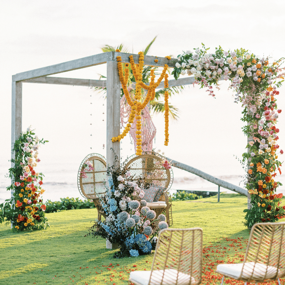 Outdoor wedding arch decorated with flowers and marigold garlands on green grass near water.