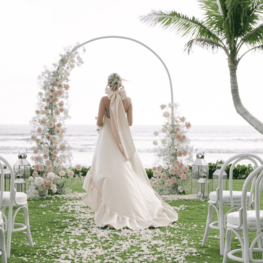 Bride in a flowing white gown with a large bow on her back walking toward a floral arch by the ocean.