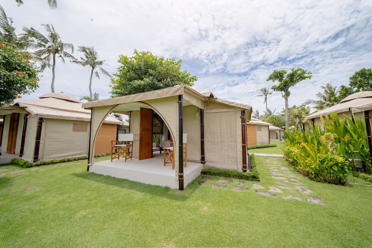 Outdoor glamping tents with wooden furniture on a wooden deck surrounded by lawn and tropical plants under a cloudy sky.