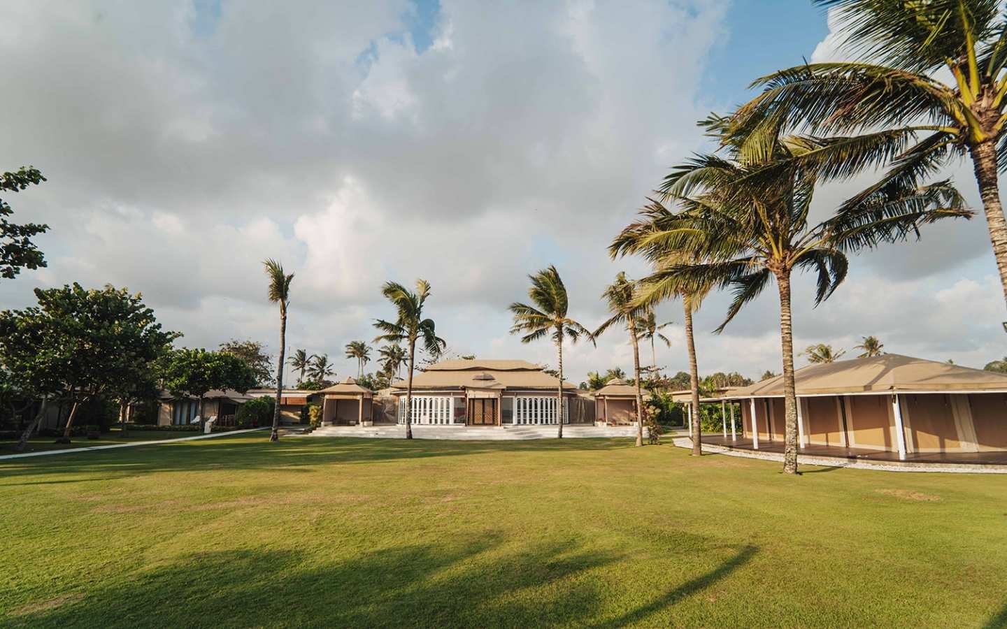 Wide green lawn with palm trees surrounding a large tropical resort building under cloudy sky.
