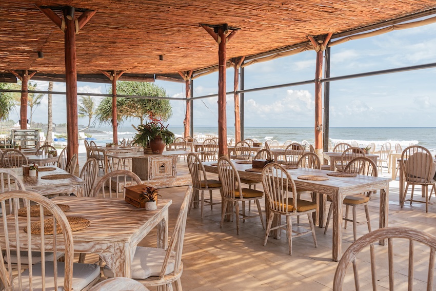 Open-air beachside restaurant with wooden tables and chairs under a thatched roof overlooking the ocean.