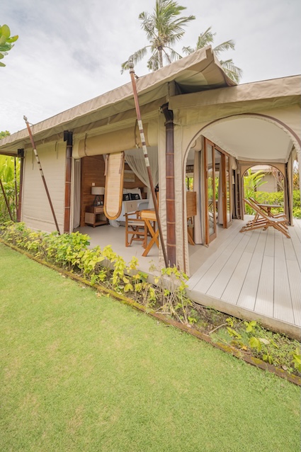Open-air luxury tent with wooden furniture and green lawn in tropical setting.