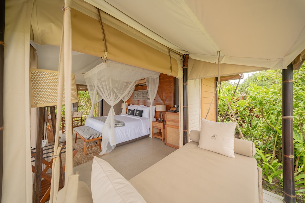 Open-air tented bedroom with a white canopy bed, wooden furniture, and a cushioned daybed surrounded by greenery.