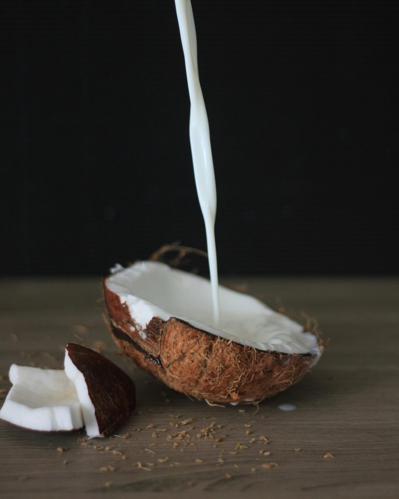 Milk being poured into a cracked open coconut half with coconut pieces on a wooden surface.