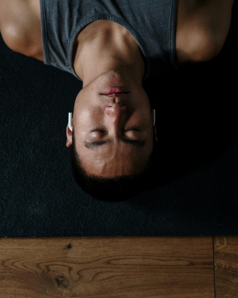 Man lying on black carpet with eyes closed, wearing gray tank top and white earbuds.