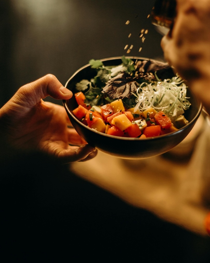 Hand holding a bowl of diced colorful vegetables and greens with seasoning being sprinkled on top.