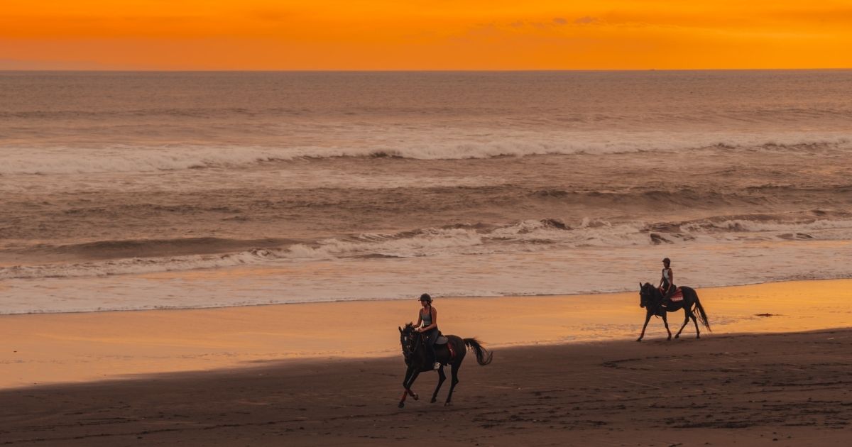 Two people riding horses on a beach at sunset with orange sky and ocean waves.