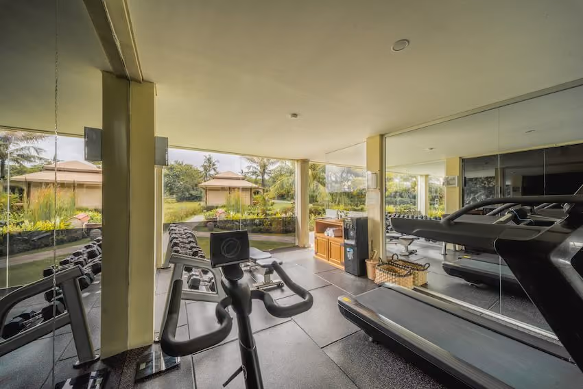 Indoor gym with a stationary bike, treadmill, dumbbells, and large windows overlooking a garden.