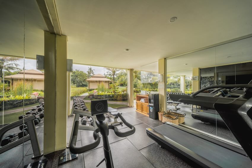 Indoor gym with a stationary bike, treadmill, dumbbells, and large windows overlooking a garden.