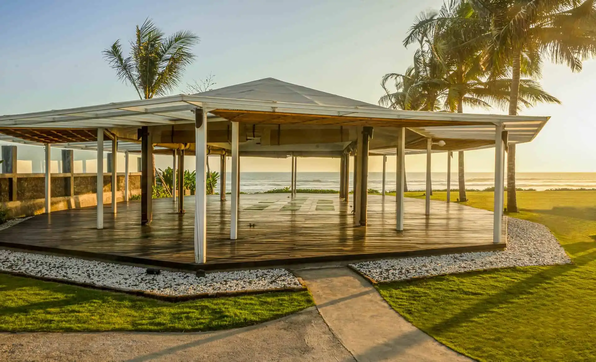 Open-air wooden pavilion with a white roof on a coastal lawn at sunset with palm trees nearby.