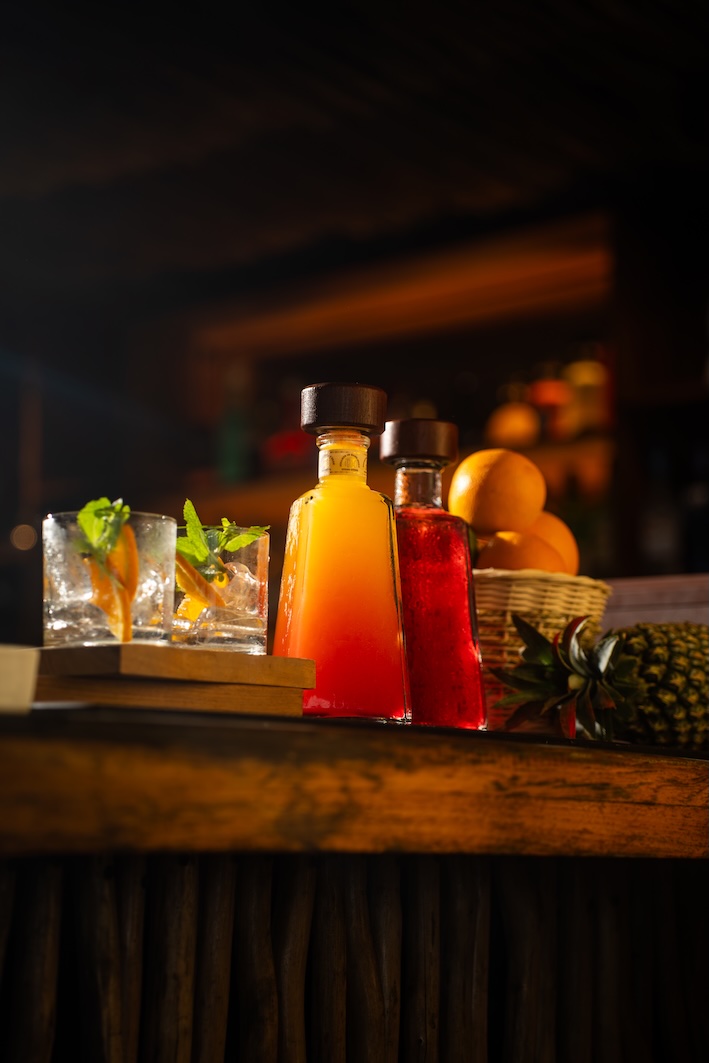 Two glass bottles with colorful liquids and two glasses with ice and mint on a wooden bar counter.