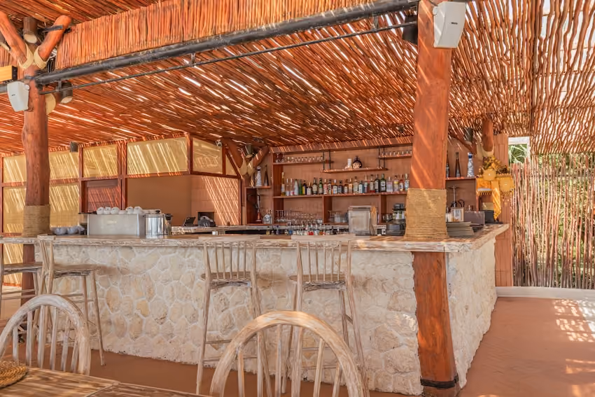 Outdoor bar with stone counter, wooden stools, and a rustic wooden roof with beams and bottles on shelves.