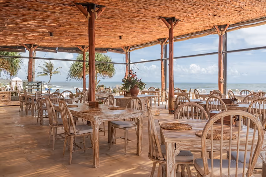 Open-air beachside restaurant with wooden tables and chairs under a thatched roof overlooking the ocean.