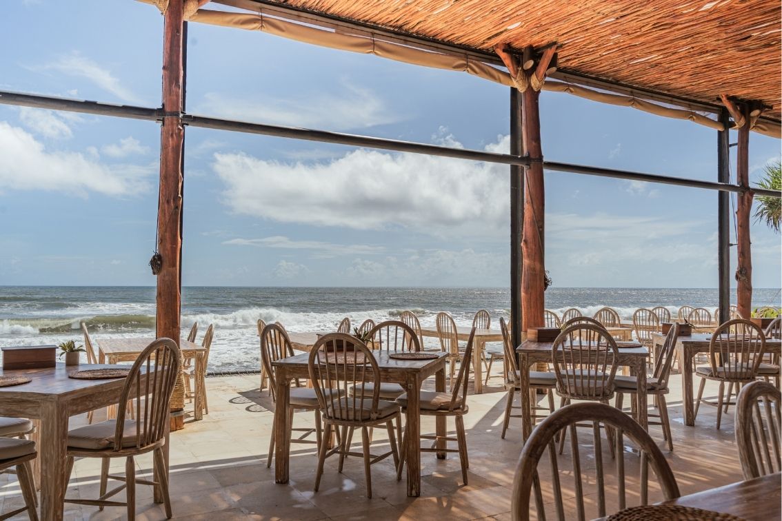 Outdoor dining area with wooden tables and chairs overlooking the ocean under a thatched roof.
