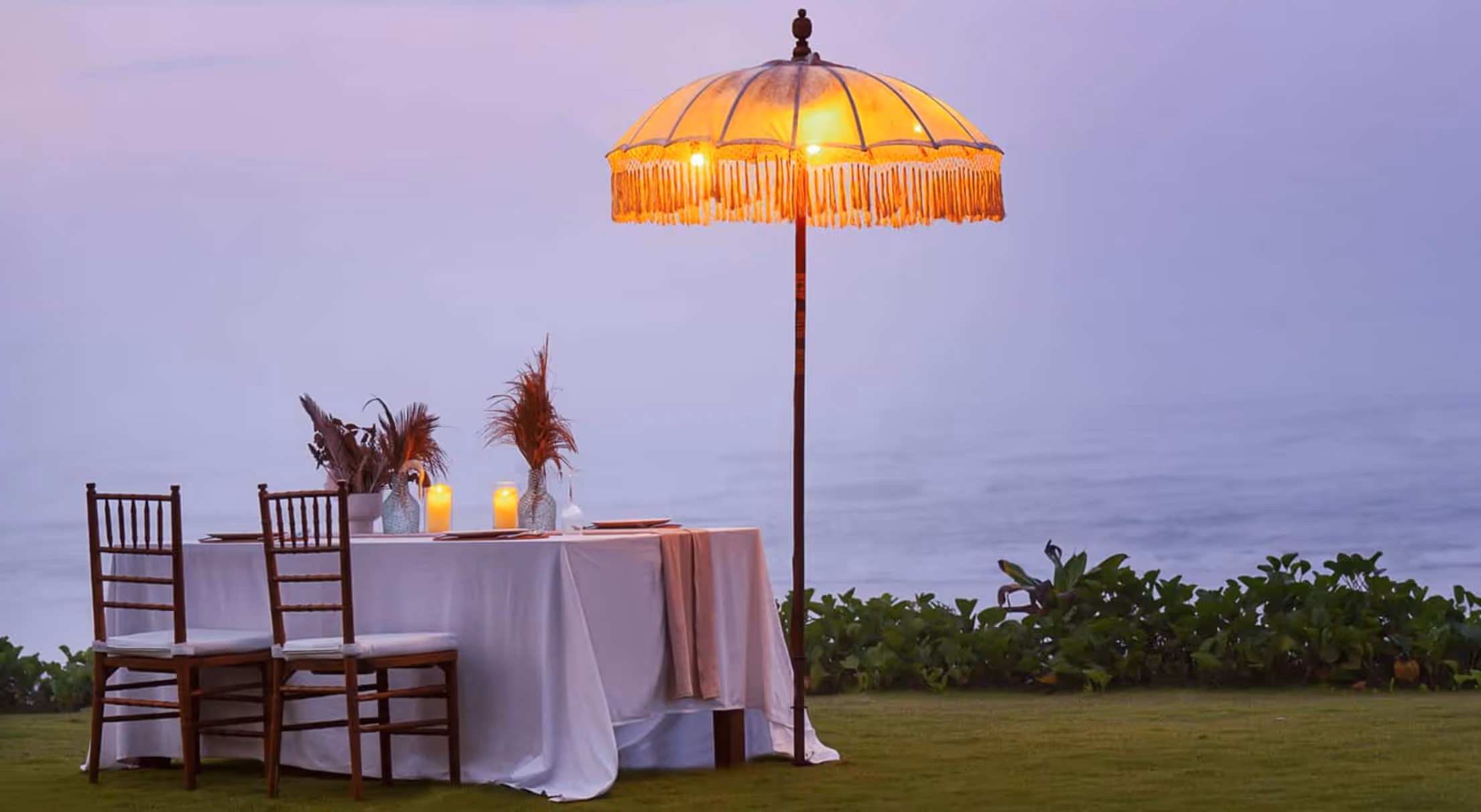 Outdoor dining table with white cloth, wooden chairs, candles, and a lit umbrella at dusk near greenery.
