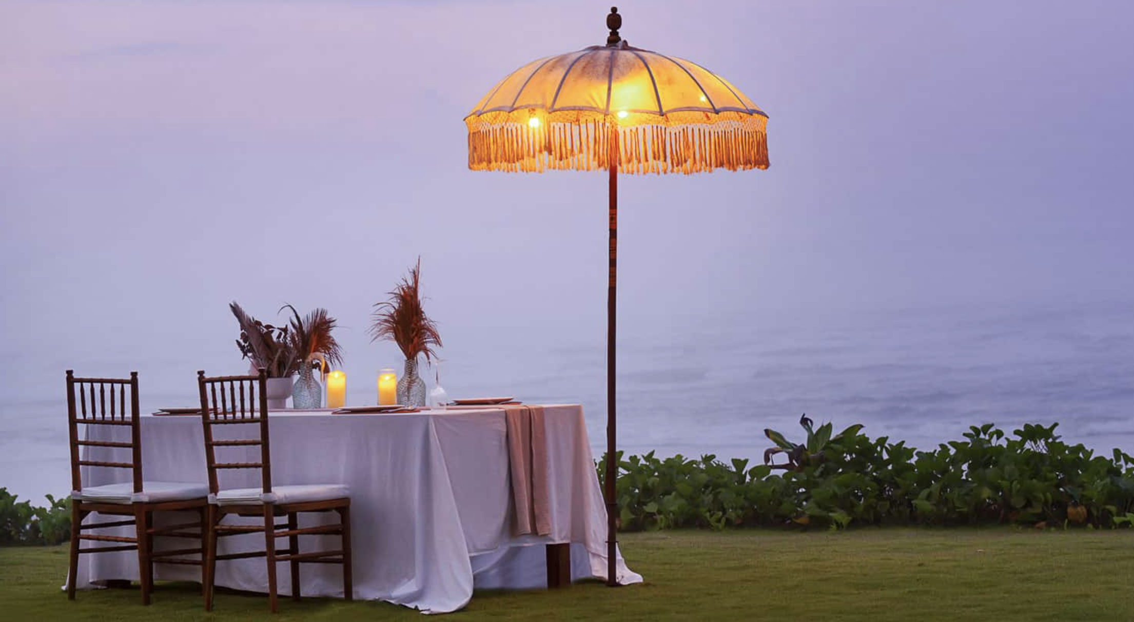 Outdoor dining table with white cloth, wooden chairs, candles, and a lit umbrella at dusk near greenery.