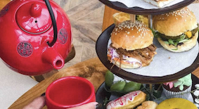 Red teapot and cup next to a tray with small burgers and sandwiches on parchment paper.