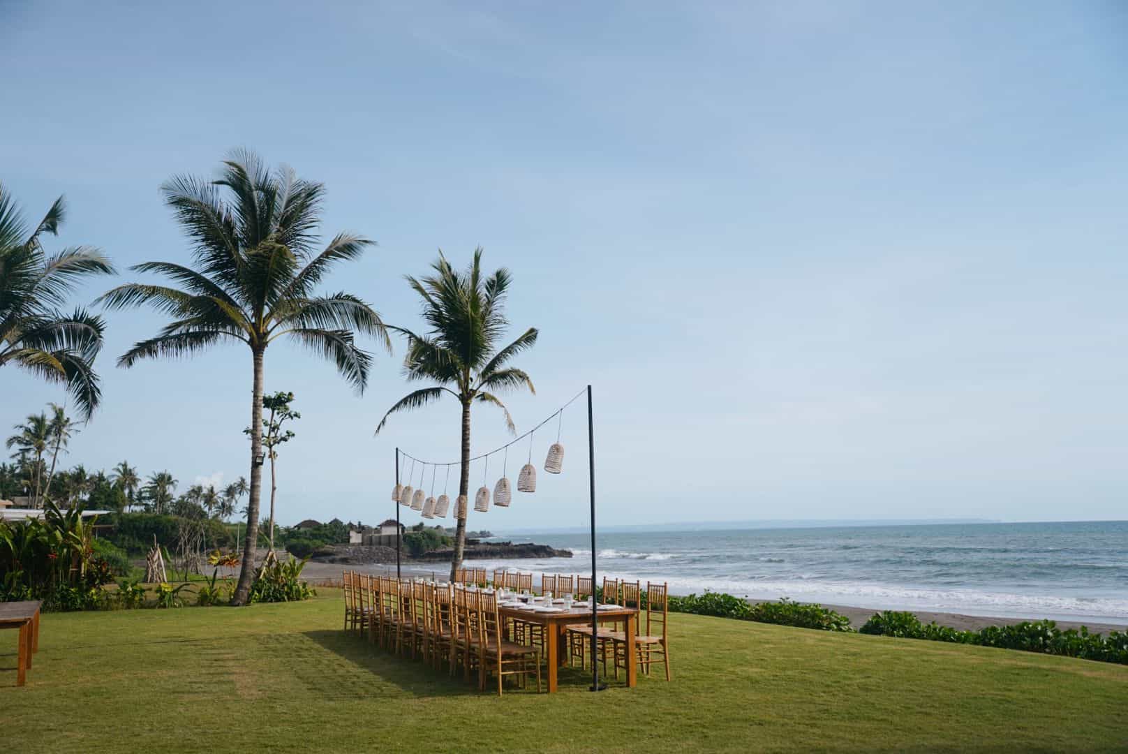Long outdoor dining table with wooden chairs set on grassy area by the ocean and palm trees.