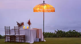 Romantic outdoor table setup with two chairs, candles, and a lit parasol by the water at dusk.