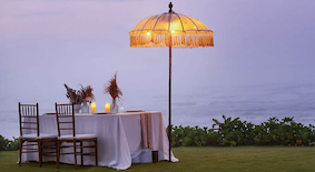Romantic outdoor table setup with two chairs, candles, and a lit parasol by the water at dusk.