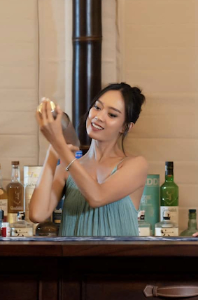 Smiling woman shaking a cocktail shaker behind a bar with bottles in the background.