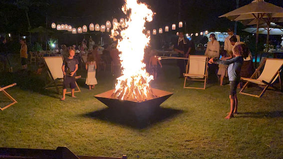 Children roasting marshmallows around a large bonfire in a grassy outdoor setting at night.