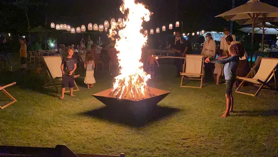 Children roasting marshmallows around a large bonfire in a grassy outdoor setting at night.