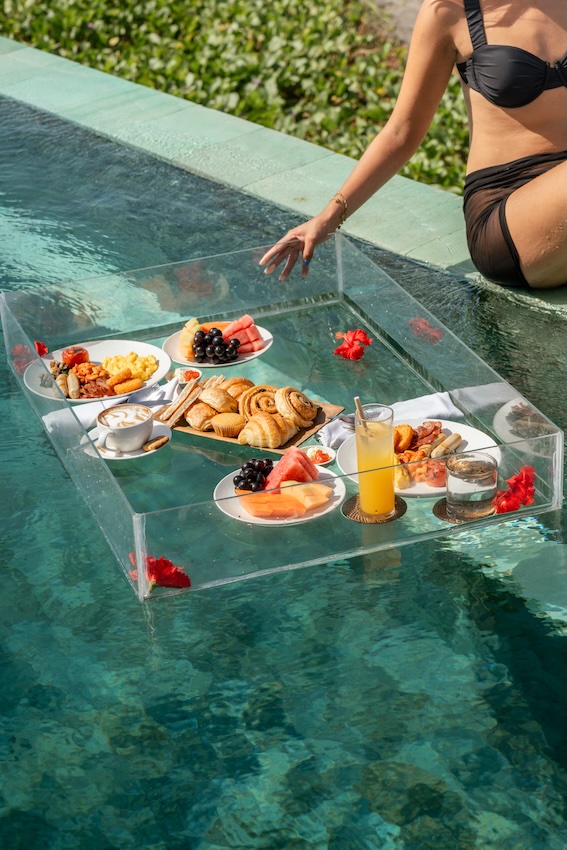 A woman in a black bikini sitting at the pool edge with a floating tray holding breakfast items and drinks in the pool.