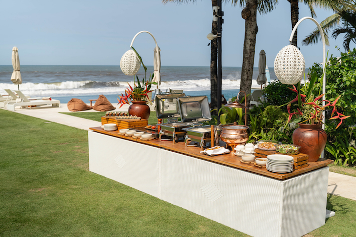 Outdoor buffet setup on a wooden counter with chafing dishes, plates, bowls, and tropical flower decorations near a beach.