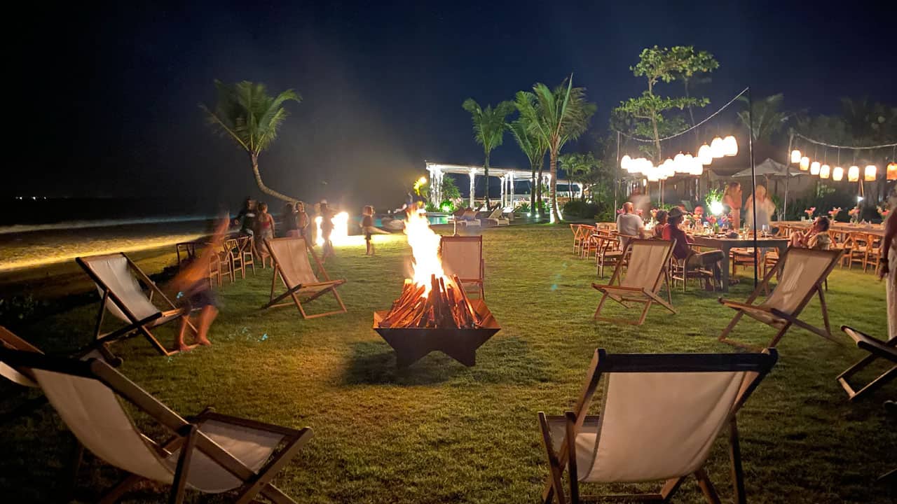 Outdoor nighttime scene with a central bonfire surrounded by empty chairs and people dining under string lights.