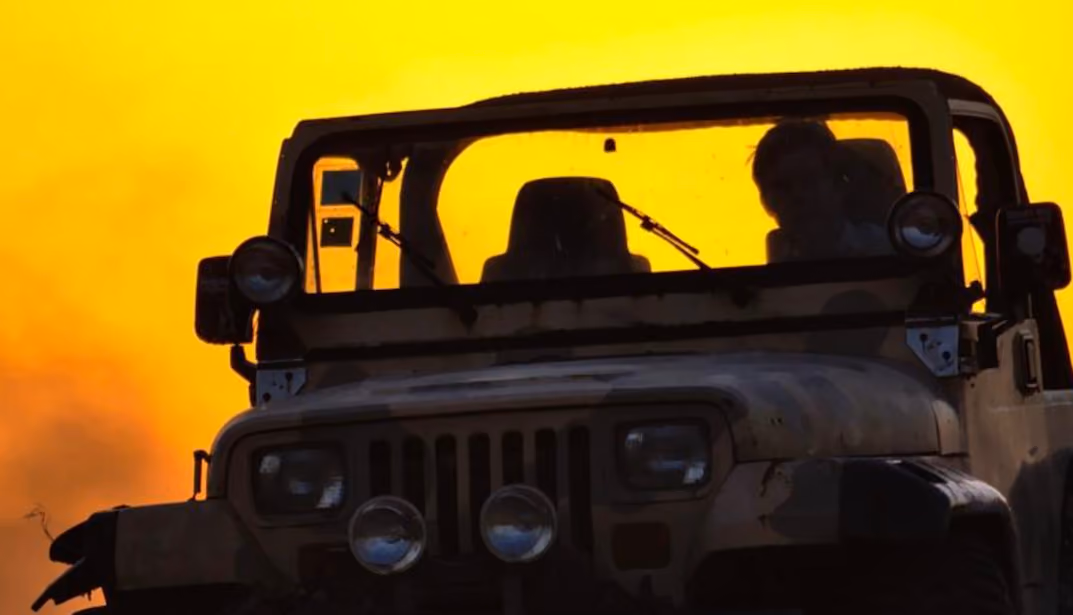 Front view of a Jeep with two people inside against a bright orange sunset sky.