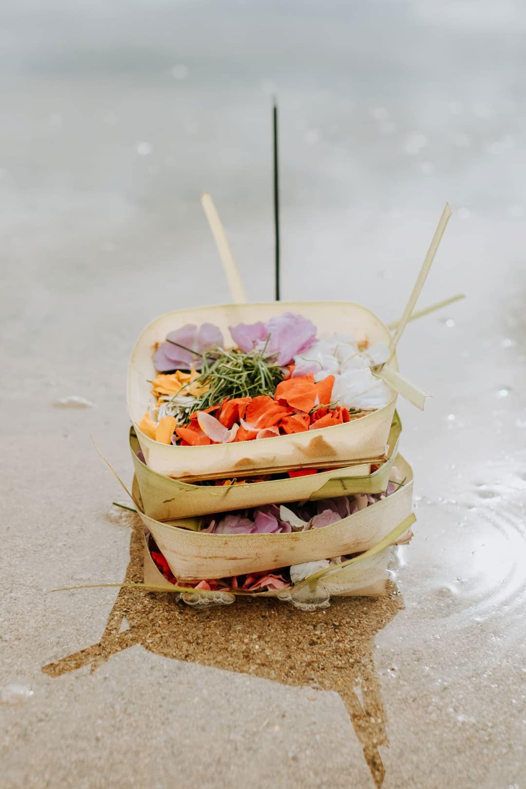 Stacked traditional Balinese flower offerings made of woven palm leaves with colorful petals on wet sand.