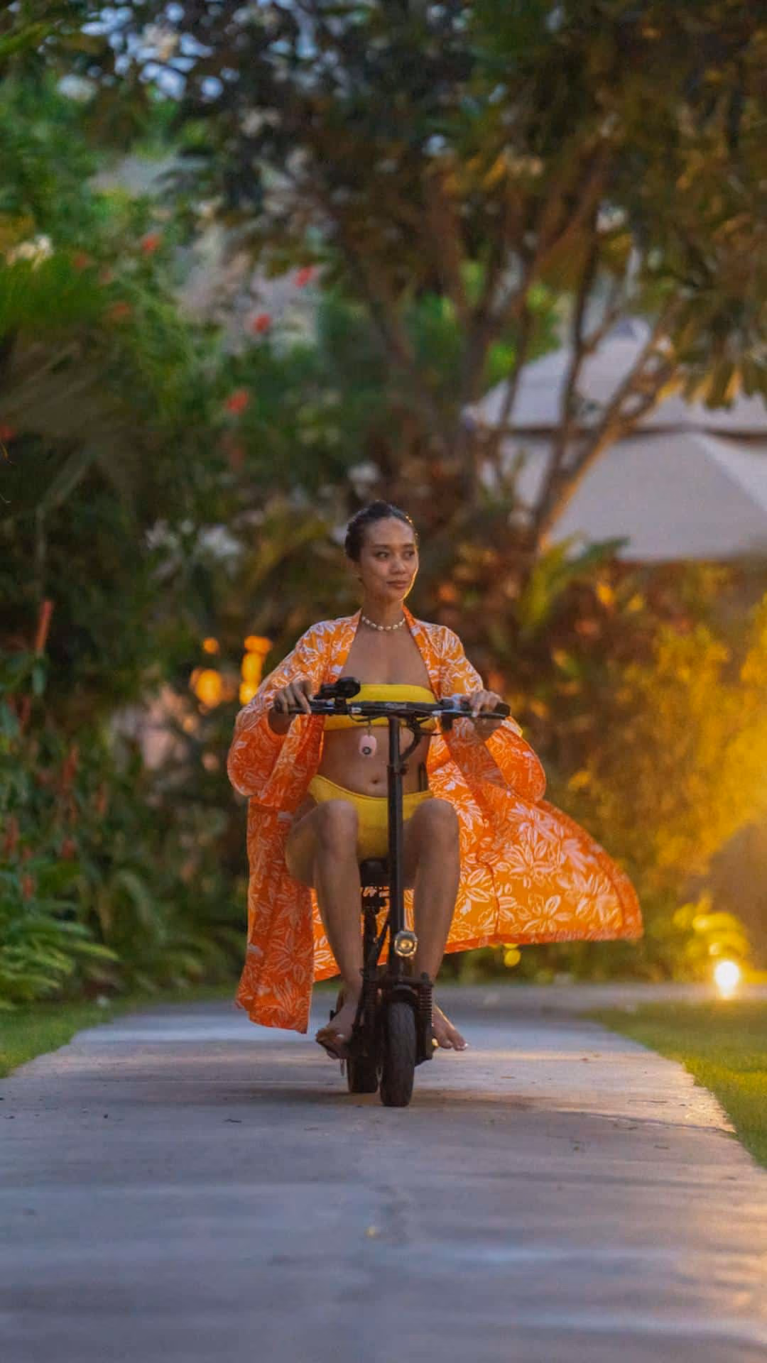 Woman in yellow swimsuit and orange cover-up riding electric scooter on garden path at dusk.
