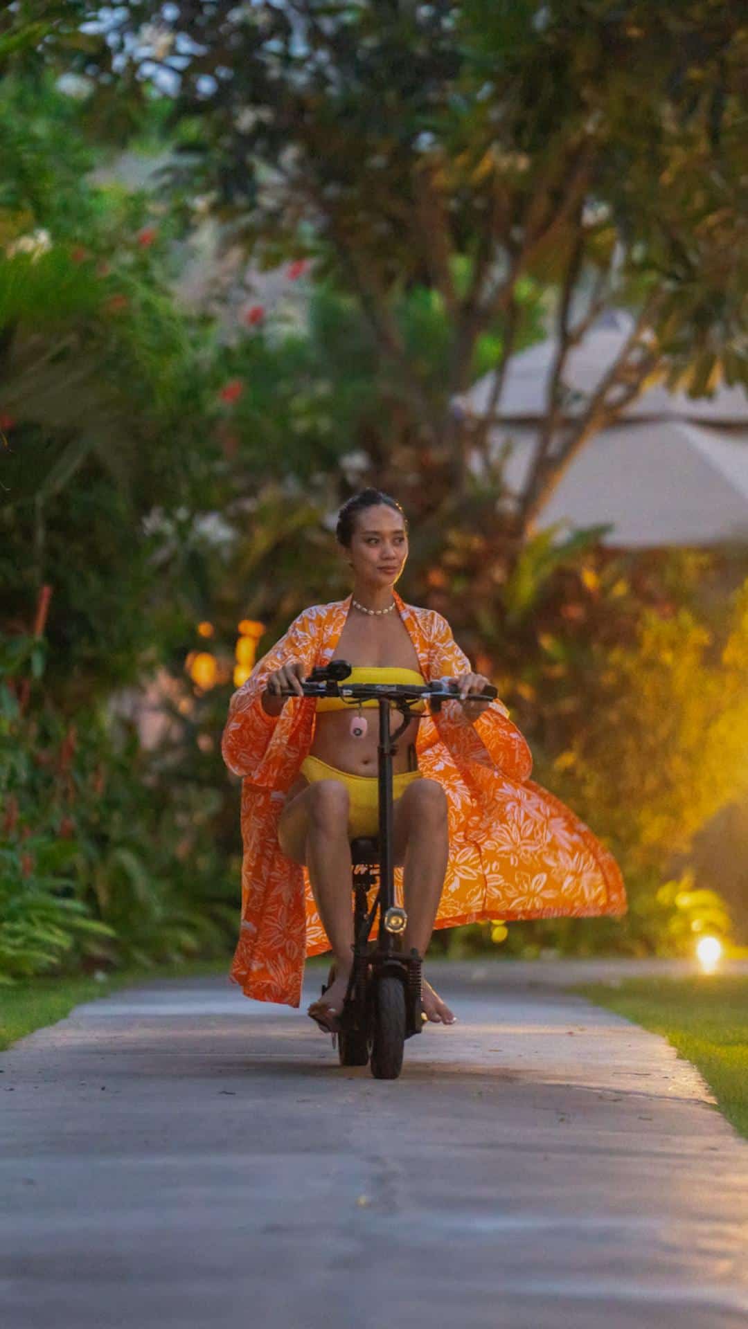Woman in yellow swimsuit and orange cover-up riding electric scooter on garden path at dusk.