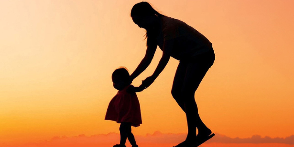 Silhouette of an adult helping a toddler walk against an orange sunset sky.