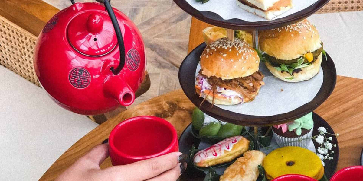 Red teapot pouring tea into a matching red cup next to a tiered tray with sliders and assorted pastries.