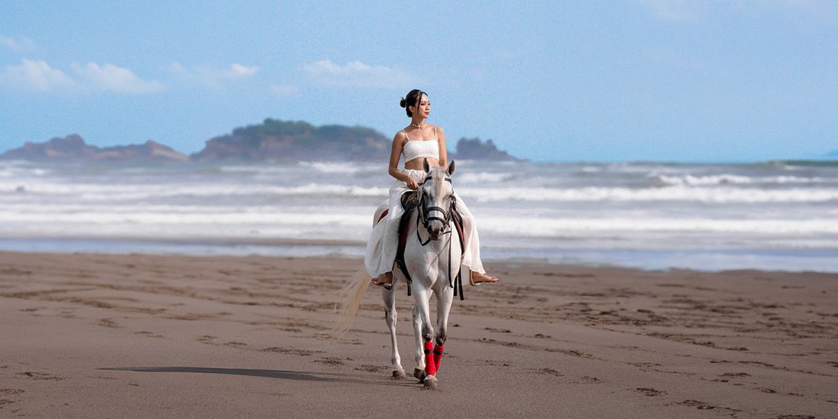 Woman in white outfit riding a white horse with red leg wraps on a sandy beach near the ocean.