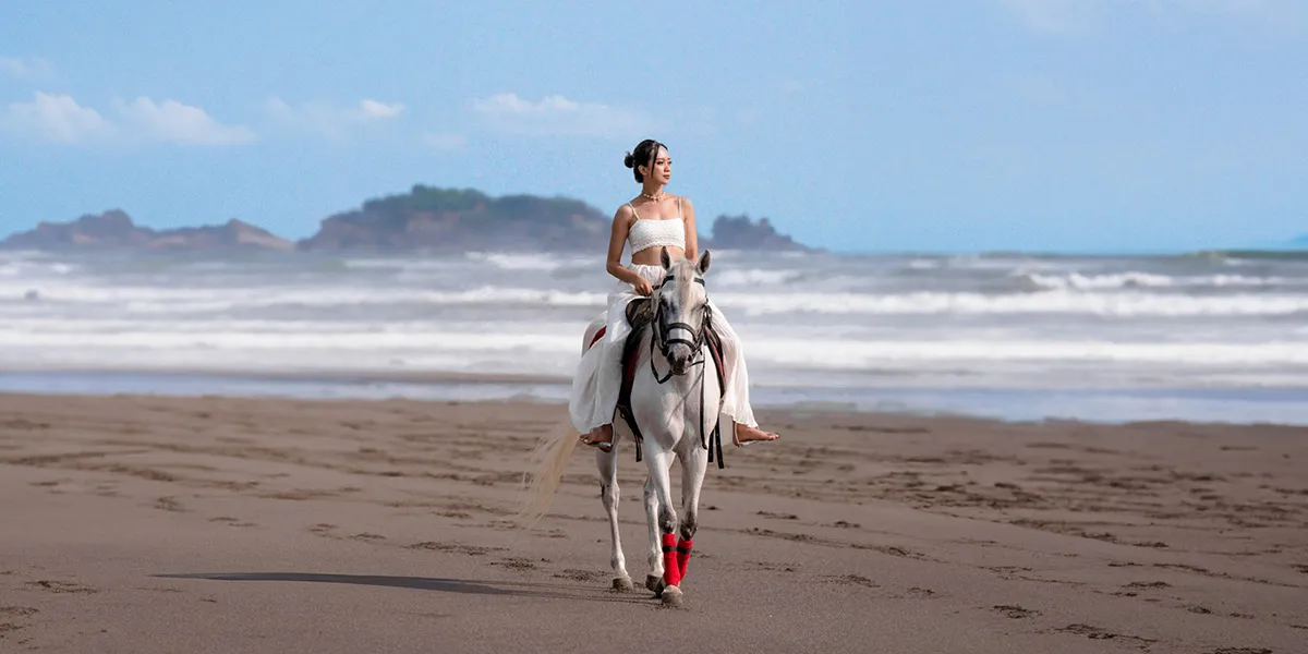 Woman in white outfit riding a white horse with red leg wraps on a sandy beach near the ocean.