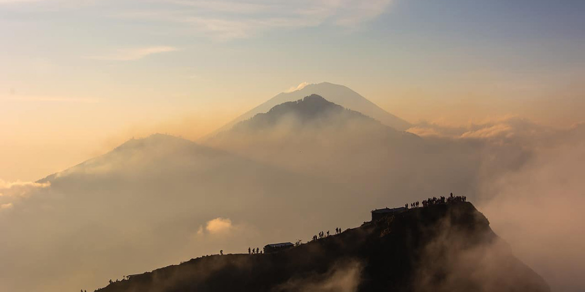 Silhouetted hikers on a foggy mountain ridge at sunrise with distant volcanic peaks in the background.