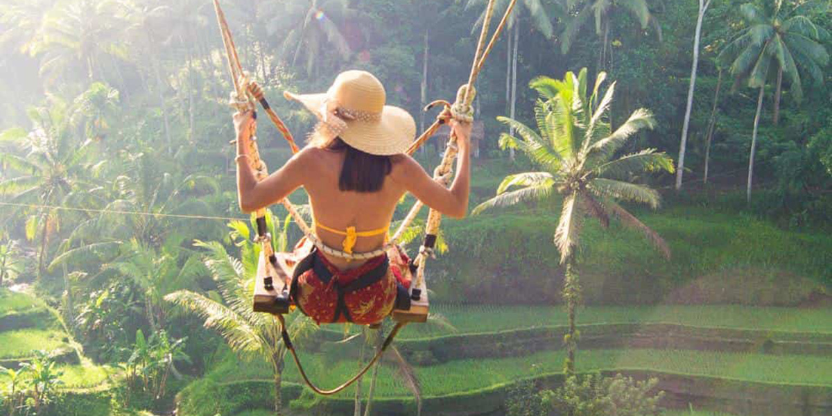 Woman wearing a sun hat swinging over lush green tropical rice terraces with palm trees around.