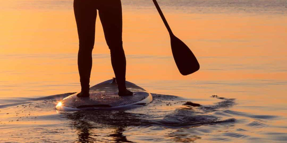 Person standing on a paddle board holding a paddle with water reflecting an orange sunset.