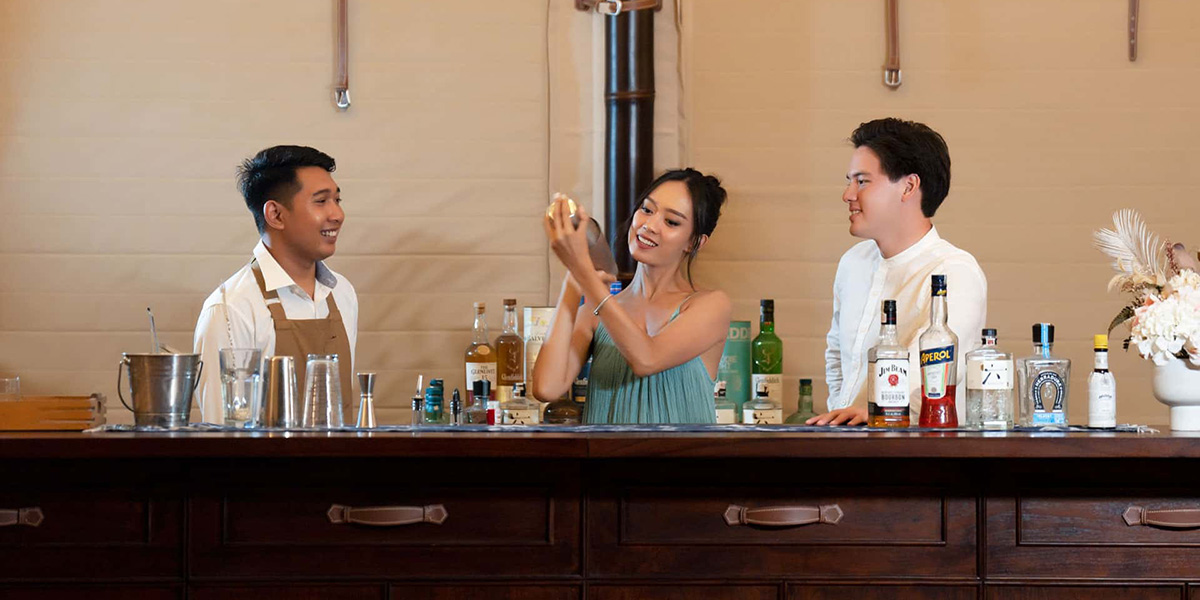 Three people behind a bar with one shaking a cocktail shaker and bottles lined up on the counter.
