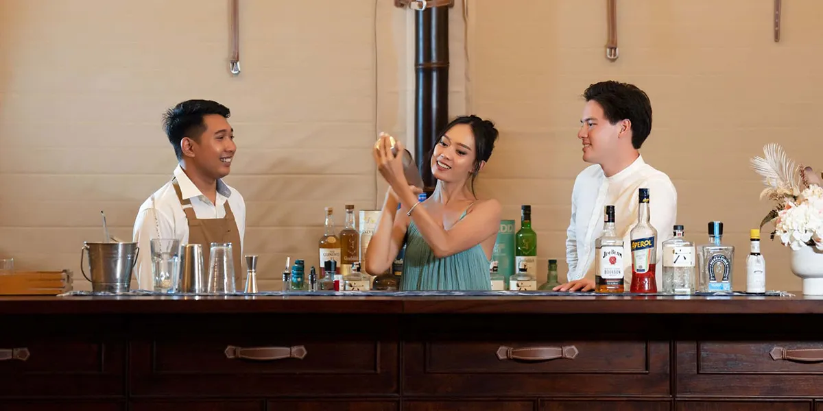 Three people behind a bar with one shaking a cocktail shaker and bottles lined up on the counter.